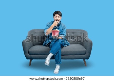 young man in a denim jacket eats popcorn from a striped bucket while watching a movie on a sofa.