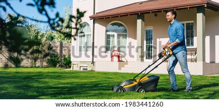 Young man in denim wear mows lawn using electric lawn mower near large country house in backyard