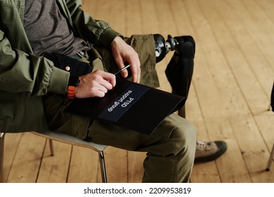 Young Man With Disability Holding Black Folder Of PTSD Support Group While Sitting On Chair During Psychological Session