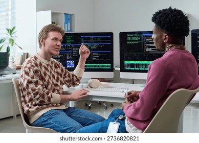 Young man discussing software development with Black young man in office, both sitting at desk with multiple computer monitors displaying code