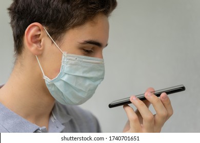 A Young Man In A Disposable Medical Protective Mask On His Face Is Talking On The Phone. Teen Hold Phone Speak On Speakerphone Using Virtual Digital Voice Recognition Assistant Search.