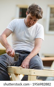 Young Man In A Diy Project Outside Hammering A Nail Into A Wooden Structure While Building A Playhouse.