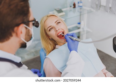 Young Man Doctor In Dentist Binocular Loupe Mask Drills Tooth Of Patient European Woman With Caries Decay Sit At Dentist Office Chair Indoor Cabinet Near Stomatologist. Healthcare Enamel Treatment.