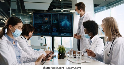 Young Man Doctor In Mask Discussing With Mixed-races Healthcare Professionals Treatment Of Coronavirus Disease In Hospital Looking At Digital Screen. Plugs Infection Covid-19. Woman Typing On Tablet