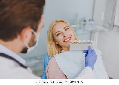 Young man doctor in white gown show veneers enamel color palette for smiling happy patient woman 20s sit at dentist office chair indoor cabinet near stomatologist. Healthcare oral aesthetic treatment. - Shutterstock ID 2034032135