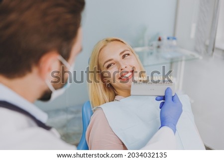 Young man doctor in white gown show veneers enamel color palette for smiling happy patient woman 20s sit at dentist office chair indoor cabinet near stomatologist. Healthcare oral aesthetic treatment.