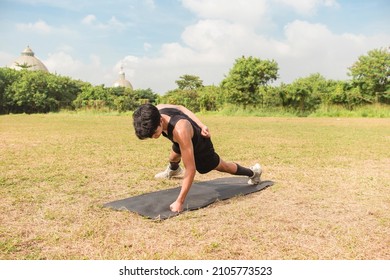 A young man does a set of one arm pushups on a black mat at an open field outdoors. Chest or full body workout.