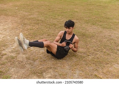 A Young Man Does A Weighted Russian Twist With A Kettlebell. An Advanced Core Workout On A Grassy Field.