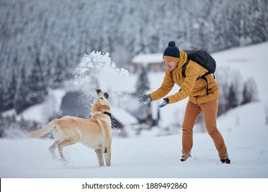 Young man with dog in winter. Pet owner with his labrador retriever playing in snow against old village in beautiful nature. Jizera mountains, Czech Republic