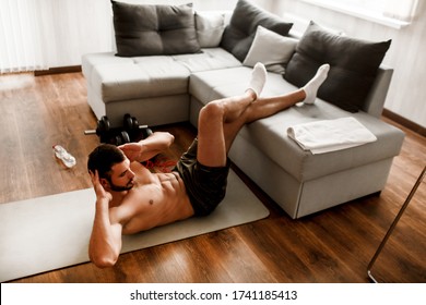 Young Man Doing Abs Exercising At Home On The Couch. Cut View Of Hardwoking T-shirtless Guy Sportsman In Workout Activity At His Apartment. Dumbbells On Pictures Lying On Floor. Trying To Get Better.