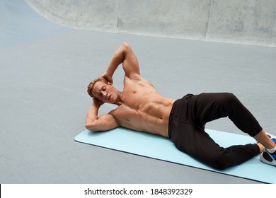 Young Man Doing Abs Workout On Yoga Mat Against Concrete Wall Outdoors. Topless Handsome Caucasian Sportsman With Strong Muscular Body In Fashion Sportswear On Functional Training. - Powered by Shutterstock