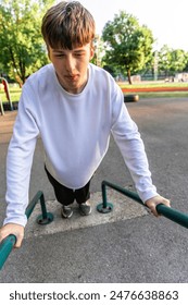 A young man is doing dips on an outdoor exercise equipment at a park. He is wearing a white shirt and black pants. The equipment is a green metal frame with two bars.