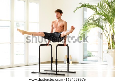 Young man doing parallel bar calisthenics exercises performing a V sit with open legs in a bright high key gym with copy space and potted palm