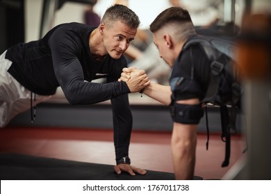 A young man is doing plank exercises with personal trainer in the gym, dressed in a black suit with an EMS electronic simulator to stimulate his muscles.