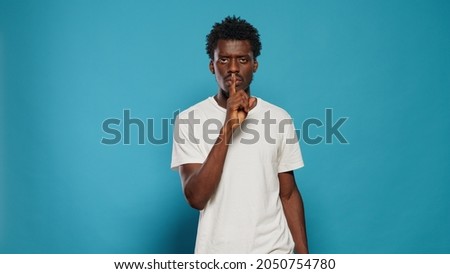 Young man doing silence gesture with finger on mouth in studio. Confident person asking for secrecy and quiet, using forefinger to make censorship expression over isolated background.