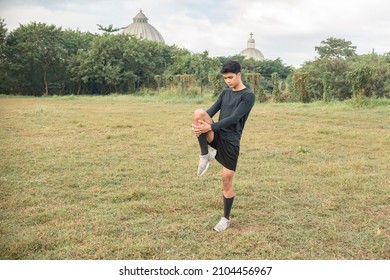 A young man doing standing glutes leg stretches before a morning run at an outdoor field.