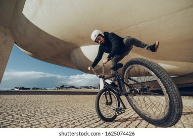 A young man is doing tricks with his BMX bicycle under a modern structure. He is wearing black clothes and white helmet. Black bmx bike. During the sunset. - Powered by Shutterstock