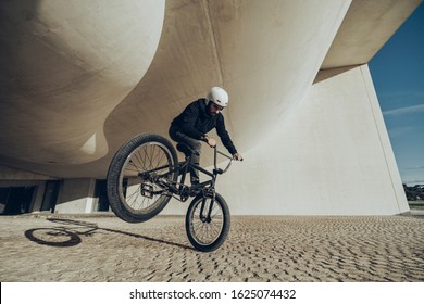 A Young Man Is Doing Tricks With His BMX Bicycle Under A Modern Structure. He Is Wearing Black Clothes And White Helmet. Black Bmx Bike. During The Sunset.