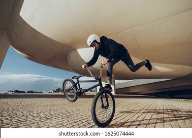 A Young Man Is Doing Tricks With His BMX Bicycle Under A Modern Structure. He Is Wearing Black Clothes And White Helmet. Black Bmx Bike. During The Sunset.