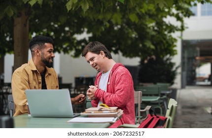 Young man with Down syndrome with his mentoring friend sitting outdoors in cafe using laptop. - Powered by Shutterstock