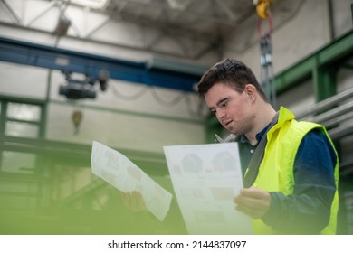 Young Man With Down Syndrome Looking At Blueprints When Working In Industrial Factory, Social Integration Concept.