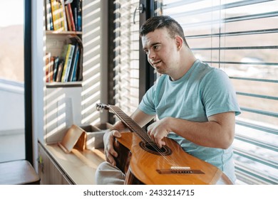 Young man with down syndrome playing acoustic guitar, sitting by window, holding and strumming guitar, making music.
