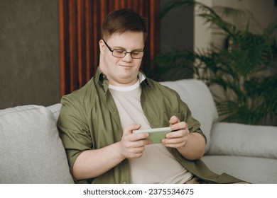 Young man with down syndrome wear casual clothes use play racing app on mobile cell phone sit on grey sofa couch stay at home flat rest spend free time in living room Genetic disease world day concept - Powered by Shutterstock