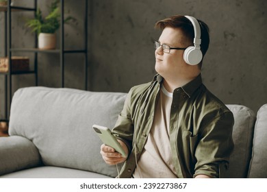 Young man with down syndrome wear clothes headphones listen music use mobile cell phone sit on grey sofa couch stay at home flat rest spend free time in living room. Genetic disease world day concept - Powered by Shutterstock