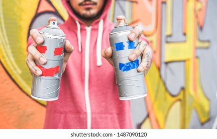 Young man drawing with two smiling sprays  - Graffiti artist painting with aerosol color cans on the wall - Urban lifestyle, youth, millennial generation and street art concept - Focus on bottles