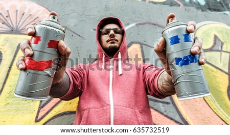 Young man drawing with two smiling sprays  - Graffiti artist painting with aerosol color cans on the wall - Rebel and against system concept - Focus on bottles spray - Unfiltered photo
