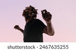 Young man with dreadlocks dances energetically in a studio, his eyes closed and arms outstretched. A pink background adds vibrancy to his silhouette as he moves to the music, using wireless earphones.