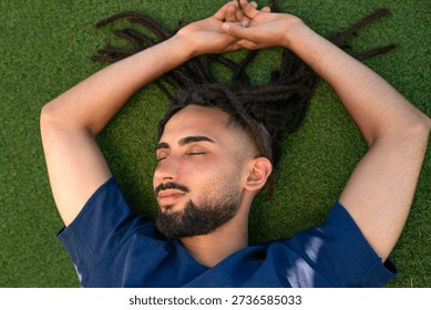 Young man with dreadlocks lying on green grass, feeling peaceful