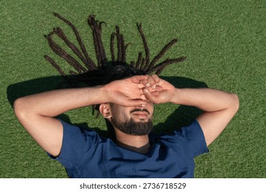 Young man with dreadlocks lying on the green grass, dazzled by the sun