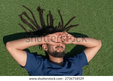 Young man with dreadlocks lying on the green grass, dazzled by the sun