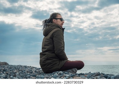 Young man with dreadlocks practicing yoga meditation is sitting in lotus position outdoors by the sea. Mental health, wellbeing, relaxing concept - Powered by Shutterstock