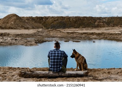 Young Man With Dreadlocks Is Sitting On Wooden Log On Riverbank With Dog Enjoying Views Of Nature. Sand Pits And Dunes And Blue Clear Water. German Shepherd With Owner. Rear View.