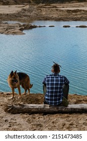 Young Man With Dreadlocks Is Sitting On Wooden Log On Riverbank With Dog Enjoying Views Of Nature. Sand Pits And Dunes And Blue Clear Water. German Shepherd With Owner. Rear View.