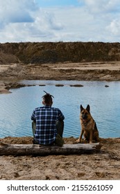 Young Man With Dreadlocks Is Sitting On Wooden Log On Riverbank With Dog Enjoying Views Of Nature. Sand Pits And Dunes And Blue Clear Water. German Shepherd With Owner. Rear View.