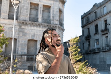 Young man with dreadlocks smiling, feeling the sun and urban lifestyle with hands on chin