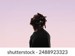 Young man with dreadlocks stands in a studio, closing his eyes in tranquility. He listens to music using wireless earbuds and a music player. His peaceful silhouette reflects his love for music. 