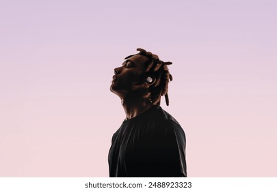 Young man with dreadlocks stands in a studio, closing his eyes in tranquility. He listens to music using wireless earbuds and a music player. His peaceful silhouette reflects his love for music. 