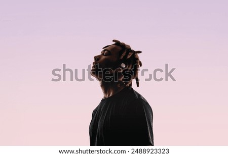 Young man with dreadlocks stands in a studio, closing his eyes in tranquility. He listens to music using wireless earbuds and a music player. His peaceful silhouette reflects his love for music. 