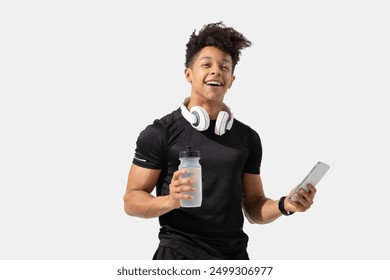 A young man dressed in black activewear stands confidently with a water bottle in one hand and a smartphone in the other. He is wearing white headphones around his neck and smiling happily - Powered by Shutterstock