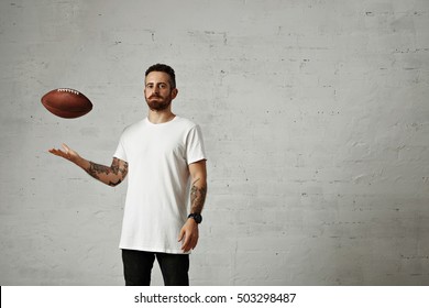 Young Man Dressed In A Blank White Cotton Sleveless T-shirt And Black Jeans Throwing A Brown Vintage Football In A Studio Isolated On White