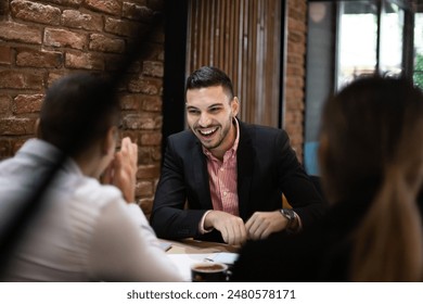 A young man, dressed in business attire, enjoys a lighthearted moment with colleagues around a cafe table, highlighting teamwork and communication. - Powered by Shutterstock