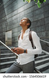 Young Man Dressed In Casual Clothes And Backpack In The City Center On The Stairs Is Resting Looking Upwards With The Smart Phone In His Hands - Millennial Is Going To Work