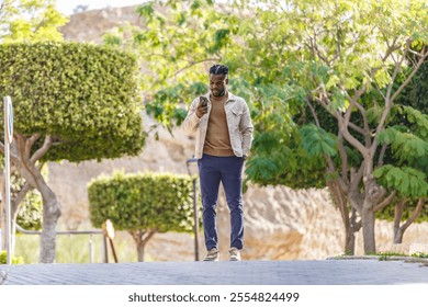 A young man dressed casually stands on a path in a park, absorbed in his smartphone. Lush greenery surrounds him, creating a serene atmosphere. - Powered by Shutterstock