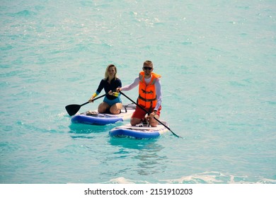 Young Man Dressed In Swimsuit And Lifevest And Young Woman Paddling On Stand Up Paddle Board. Summer Outdoor Seaside Activities