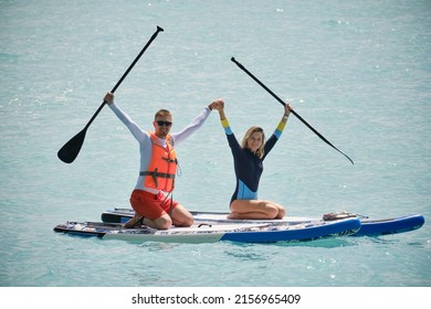 Young Man Dressed In Swimsuit And Lifevest And Young Woman Holding Hands On Stand Up Paddle Board. Summer Outdoor Seaside Activities. Family Outdoor Summer Activities