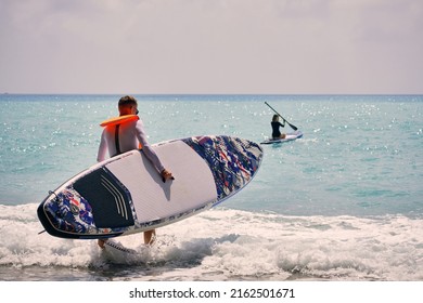 Young Man Dressed In Swimsuit And Lifevest Carrying Sup Board Towards Sea. Young Woman Paddling On Stand Up Paddle Board At Background. Summer Outdoor Seaside Activities